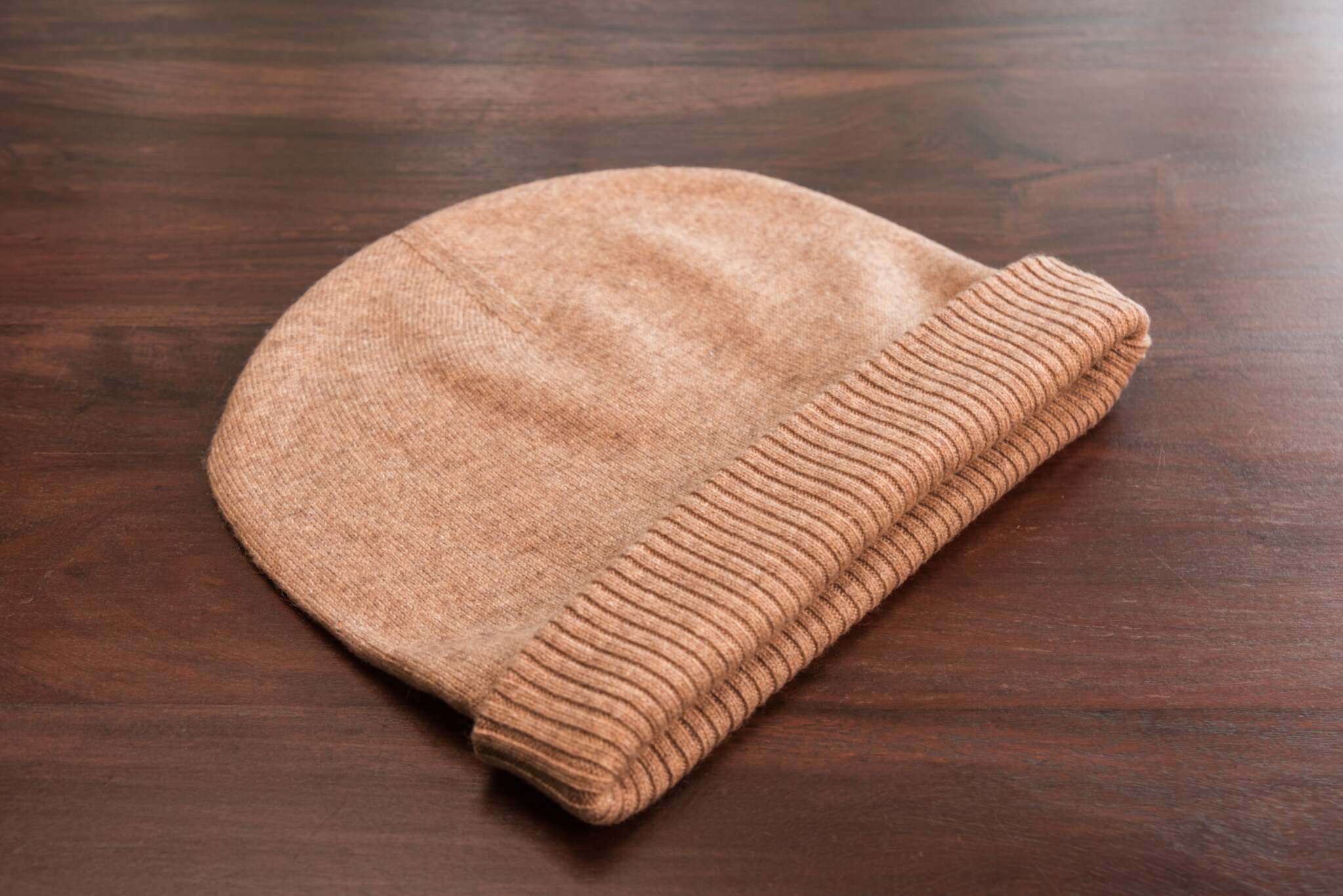 Auf dem Bild ist eine braune Beanie-Mütze aus feinem, vermutlich kaschmirartigem Material zu sehen. Die Mütze liegt flach auf einer dunklen Holzoberfläche. Sie hat eine glatte Struktur und ist am unteren Rand mit einem gerippten Umschlag versehen. Die Farben wirken weich und warm, und das Material sieht sehr weich und hochwertig aus. Es sind keine weiteren Objekte oder Dekorationen im Bild sichtbar, der Fokus liegt ganz auf der Mütze.