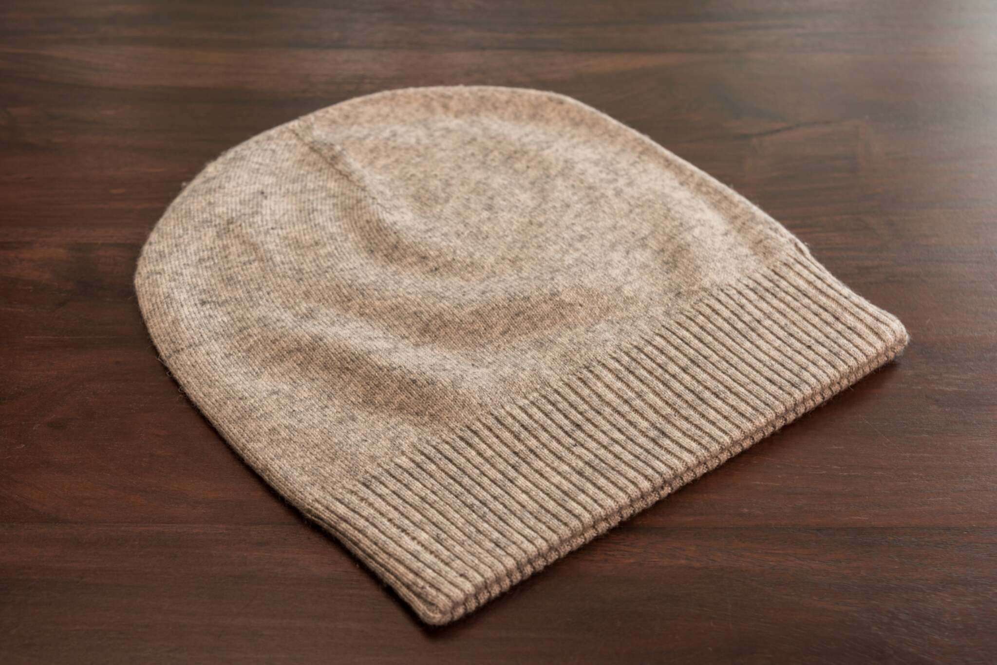 Auf dem Bild ist eine hellbraune Strickmütze aus weichem Material zu sehen, mit einem gerippten, umgeschlagenen Bund. Die Mütze liegt flach auf einer glatten, dunkelbraunen Holzoberfläche. Es sind keine weiteren Gegenstände oder auffälligen Merkmale im Hintergrund zu erkennen. Die Textur der Mütze wirkt flauschig und gleichmäßig gestrickt.