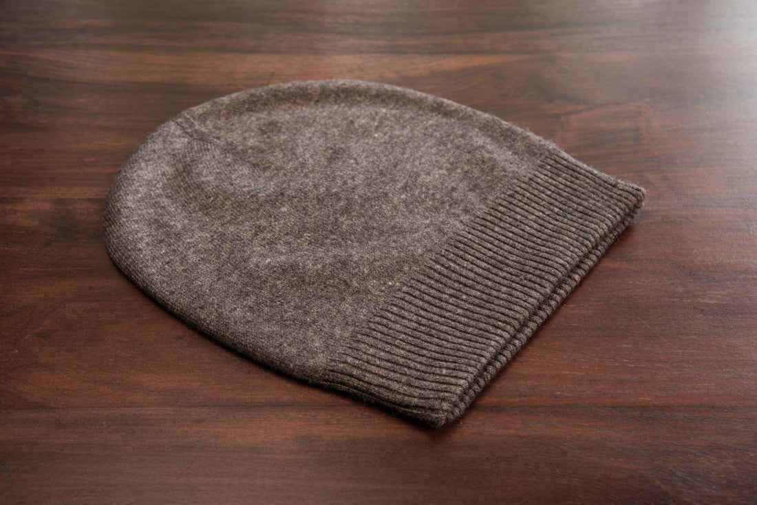 Auf dem Bild ist eine dunkelgraue Kaschmirmütze zu sehen, die flach auf einer glatten, braunen Holzoberfläche liegt. Die Mütze hat ein schlichtes, glattes Strickmuster und einen breiten, gerippten Umschlag am unteren Rand. Das Material wirkt weich und hochwertig. Sonstige Objekte oder Dekorationen sind nicht sichtbar. Die Beleuchtung ist natürlich und gleichmäßig, wodurch die Struktur und Farbe der Mütze gut erkennbar sind.