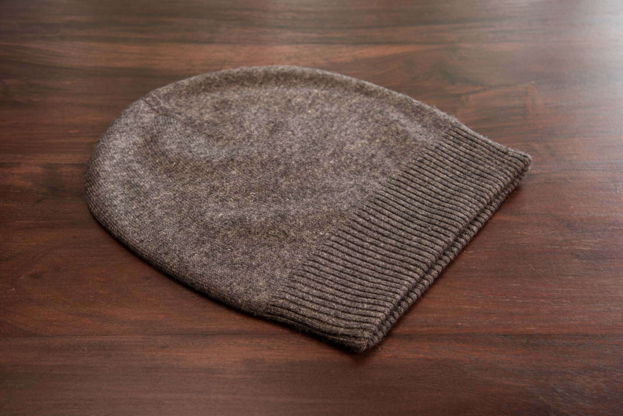 Auf dem Bild ist eine dunkelgraue Kaschmirmütze zu sehen, die flach auf einer glatten, braunen Holzoberfläche liegt. Die Mütze hat ein schlichtes, glattes Strickmuster und einen breiten, gerippten Umschlag am unteren Rand. Das Material wirkt weich und hochwertig. Sonstige Objekte oder Dekorationen sind nicht sichtbar. Die Beleuchtung ist natürlich und gleichmäßig, wodurch die Struktur und Farbe der Mütze gut erkennbar sind.