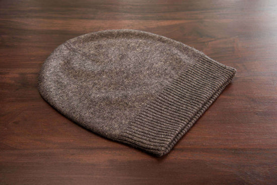 Auf dem Bild ist eine dunkelgraue Kaschmirmütze zu sehen, die flach auf einer glatten, braunen Holzoberfläche liegt. Die Mütze hat ein schlichtes, glattes Strickmuster und einen breiten, gerippten Umschlag am unteren Rand. Das Material wirkt weich und hochwertig. Sonstige Objekte oder Dekorationen sind nicht sichtbar. Die Beleuchtung ist natürlich und gleichmäßig, wodurch die Struktur und Farbe der Mütze gut erkennbar sind.