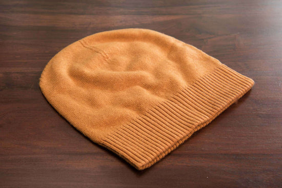 Auf dem Bild ist eine hellbraune, gestrickte Beanie-Mütze aus weichem Stoff zu sehen. Die Mütze hat einen breiten, gerippten Umschlag am unteren Rand. Sie liegt flach auf einer dunkelbraunen, hölzernen Oberfläche. Das Strickmuster ist glatt, nur der Umschlag ist deutlich gerippt und hebt sich optisch ab. Die Mütze wirkt gemütlich und dehnbar, gut geeignet für kühle Tage. Weitere Gegenstände sind nicht zu erkennen; das Bild konzentriert sich auf die Mütze.