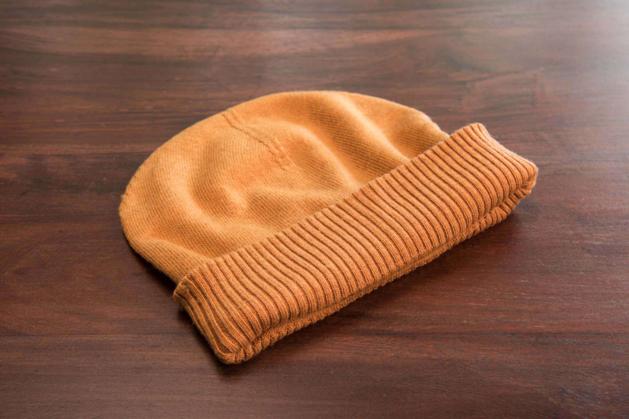 Auf dem Bild ist eine hellbraune, gestrickte Beanie-Mütze mit glatter Oberfläche und einem gerippten Umschlag zu sehen. Die Mütze liegt flach auf einer glatten Holzoberfläche. Es sind keine weiteren Gegenstände oder Muster im Hintergrund erkennbar. Die Mütze wirkt weich und hochwertig verarbeitet.
