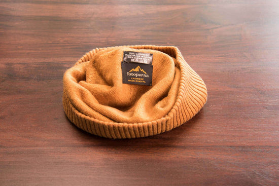 Auf dem Bild ist eine hellbraune Kaschmirmütze mit glatt gestricktem Hauptteil und geripptem Umschlag zu erkennen. Die Mütze liegt auf einer Holzoberfläche. Im Inneren der Mütze sind zwei schwarze Etiketten sichtbar: Auf einem steht „100% Cashmere, Handmade in Nepal, Dry Clean Only“, auf dem anderen befindet sich das Logo „YETOPURNA Cashmere Made in Nepal“ in goldener Schrift. Die Mütze wirkt weich und hochwertig verarbeitet.