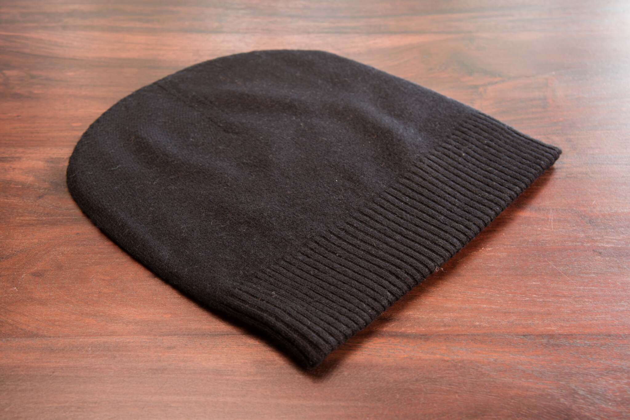 Auf dem Bild liegt eine schwarze Kaschmirmütze im Beanie-Stil auf einer glatten, braunen Holzoberfläche. Der Mützenumschlag ist gerippt gestrickt, der Rest der Mütze erscheint glatt. Es sind keine weiteren Gegenstände sichtbar.