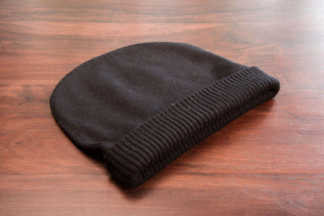 Auf dem Bild ist eine schwarze Mütze aus feinem Strick zu sehen, die auf einer braunen, hölzernen Oberfläche liegt. Der Umschlag der Mütze ist gerippt und hebt sich optisch gut vom glatt gestrickten Hauptteil ab. Das Material wirkt weich und leicht flauschig. Sonst sind keine weiteren Gegenstände auf dem Bild.