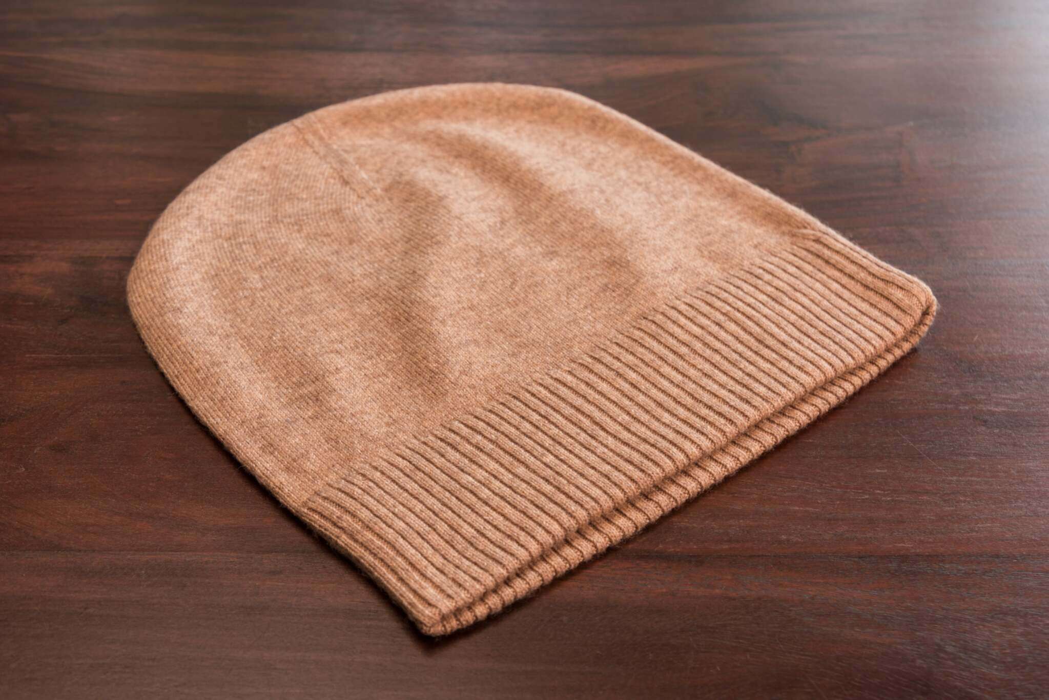 Auf dem Bild ist eine braune Beanie-Mütze aus Kaschmir zu sehen, glatt gestrickt mit einem gerippten Umschlag, liegend auf einer dunklen Holzoberfläche. Die Mütze wirkt weich und hochwertig verarbeitet. Es gibt keine weiteren sichtbaren Gegenstände im Bild.