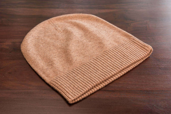 Auf dem Bild ist eine braune Beanie-Mütze aus Kaschmir zu sehen, glatt gestrickt mit einem gerippten Umschlag, liegend auf einer dunklen Holzoberfläche. Die Mütze wirkt weich und hochwertig verarbeitet. Es gibt keine weiteren sichtbaren Gegenstände im Bild.
