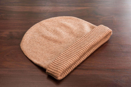 Auf dem Bild ist eine braune Beanie-Mütze aus feinem, vermutlich kaschmirartigem Material zu sehen. Die Mütze liegt flach auf einer dunklen Holzoberfläche. Sie hat eine glatte Struktur und ist am unteren Rand mit einem gerippten Umschlag versehen. Die Farben wirken weich und warm, und das Material sieht sehr weich und hochwertig aus. Es sind keine weiteren Objekte oder Dekorationen im Bild sichtbar, der Fokus liegt ganz auf der Mütze.