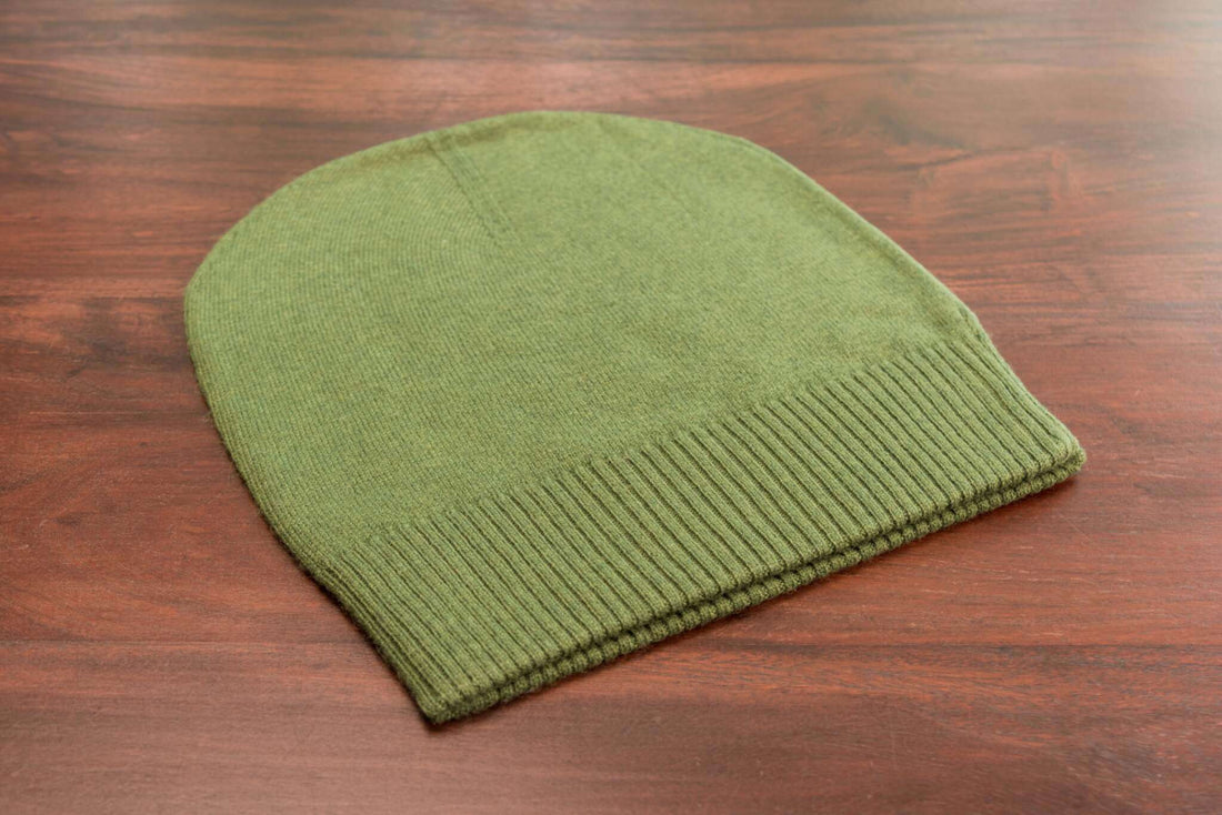 Auf dem Bild ist eine glatt gestrickte, dunkelgrüne Kaschmirmütze im Beanie-Stil zu sehen. Die Mütze liegt flach auf einer Holzoberfläche. Der untere Bereich der Mütze besitzt einen gerippten Umschlag, der sich farblich und textural leicht vom Rest unterscheidet. Das Strickmuster ist gleichmäßig und fein. Die Oberfläche wirkt weich und hochwertig. Sonst sind keine weiteren Gegenstände oder Deko-Elemente erkennbar. Die Szene ist gut ausgeleuchtet, sodass die Struktur und Farbe der Mütze klar sichtbar sind.