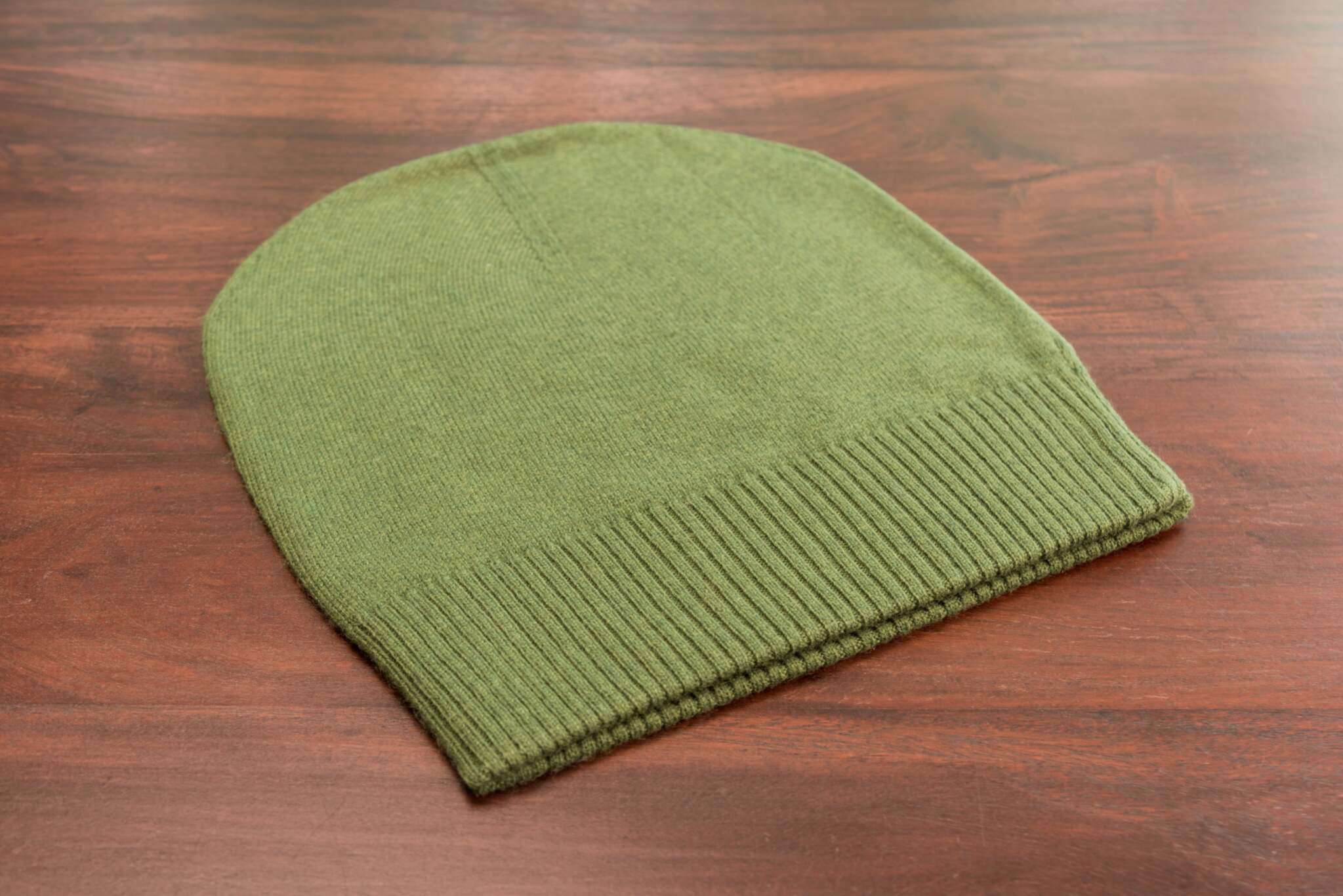 Auf dem Bild ist eine glatt gestrickte, dunkelgrüne Kaschmirmütze im Beanie-Stil zu sehen. Die Mütze liegt flach auf einer Holzoberfläche. Der untere Bereich der Mütze besitzt einen gerippten Umschlag, der sich farblich und textural leicht vom Rest unterscheidet. Das Strickmuster ist gleichmäßig und fein. Die Oberfläche wirkt weich und hochwertig. Sonst sind keine weiteren Gegenstände oder Deko-Elemente erkennbar. Die Szene ist gut ausgeleuchtet, sodass die Struktur und Farbe der Mütze klar sichtbar sind.