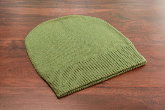 Auf dem Bild ist eine glatt gestrickte, dunkelgrüne Kaschmirmütze im Beanie-Stil zu sehen. Die Mütze liegt flach auf einer Holzoberfläche. Der untere Bereich der Mütze besitzt einen gerippten Umschlag, der sich farblich und textural leicht vom Rest unterscheidet. Das Strickmuster ist gleichmäßig und fein. Die Oberfläche wirkt weich und hochwertig. Sonst sind keine weiteren Gegenstände oder Deko-Elemente erkennbar. Die Szene ist gut ausgeleuchtet, sodass die Struktur und Farbe der Mütze klar sichtbar sind.