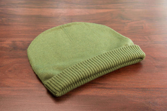 Auf dem Bild liegt eine dunkelgrüne, glatt gestrickte Beanie-Mütze mit einem gerippten Umschlag auf einer braunen Holzoberfläche. Die Mütze ist ordentlich zusammengelegt und wirkt weich und hochwertig. Im Hintergrund ist nichts anderes erkennbar, der Fokus liegt komplett auf der Mütze.