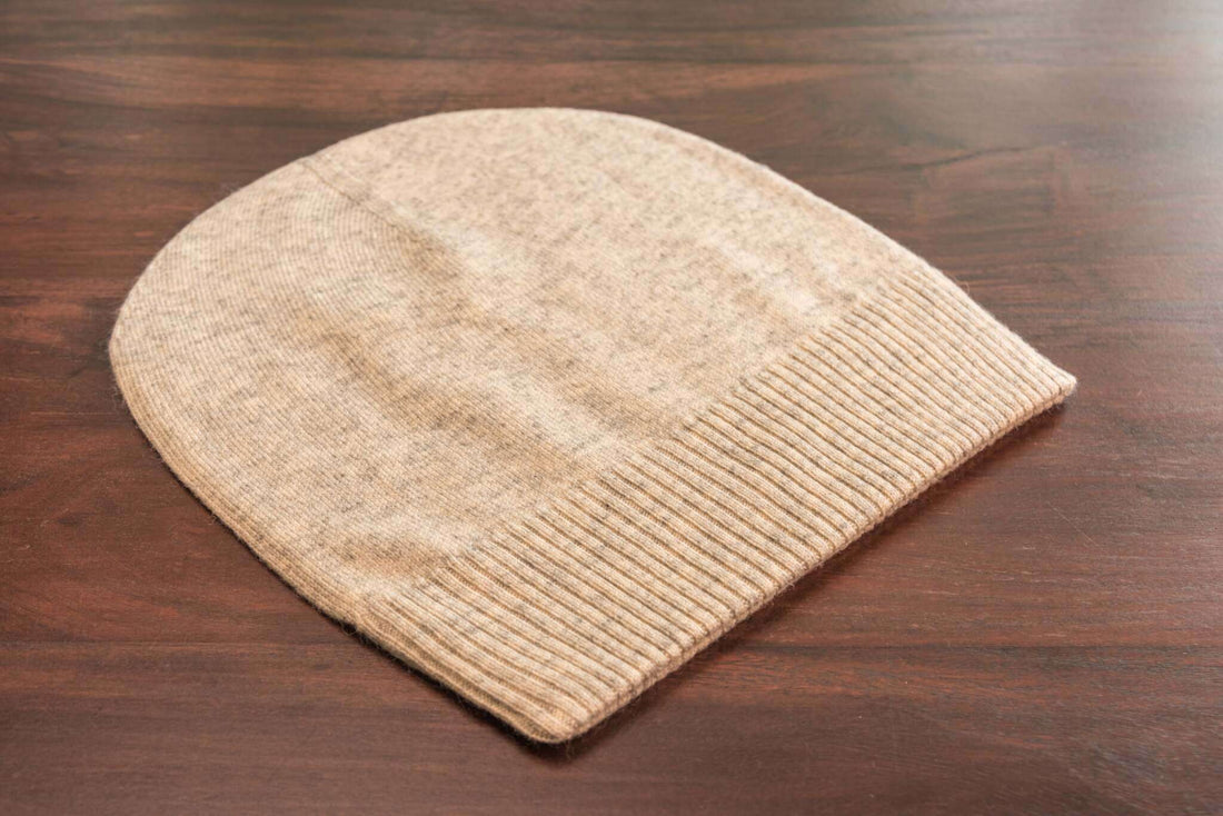 Auf dem Bild ist eine beige gestrickte Mütze aus weichem Material zu sehen, die flach auf einer dunklen Holzoberfläche liegt. Die Mütze hat einen breiten, gerippten Umschlag am unteren Rand, während der obere Teil glatt gestrickt ist. Die Farbe wirkt hellbeige oder naturfarben und das Material sieht flauschig und gemütlich aus. Außer der Mütze und der Holzoberfläche sind keine weiteren Gegenstände oder Details erkennbar.