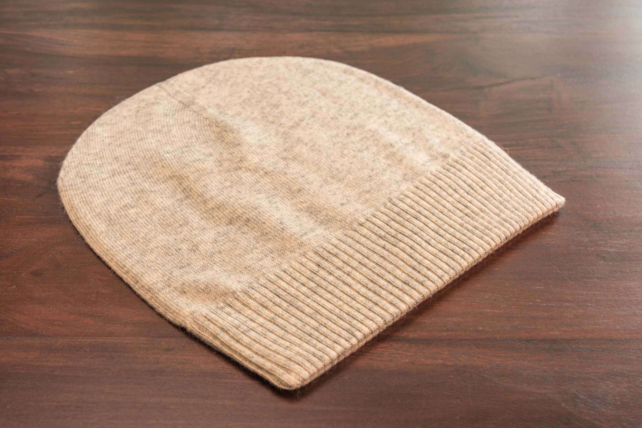 Auf dem Bild ist eine beige gestrickte Mütze aus weichem Material zu sehen, die flach auf einer dunklen Holzoberfläche liegt. Die Mütze hat einen breiten, gerippten Umschlag am unteren Rand, während der obere Teil glatt gestrickt ist. Die Farbe wirkt hellbeige oder naturfarben und das Material sieht flauschig und gemütlich aus. Außer der Mütze und der Holzoberfläche sind keine weiteren Gegenstände oder Details erkennbar.