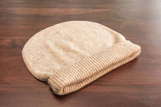Auf dem Bild liegt eine beige-weiße Strickmütze mit einem breiten, gerippten Umschlag auf einer glatten, dunklen Holzfläche. Die Mütze wirkt weich, ist glatt gestrickt und scheint aus einem feinen Material wie Kaschmir oder Wolle zu bestehen. Es sind keine weiteren Gegenstände oder Dekorationen sichtbar.