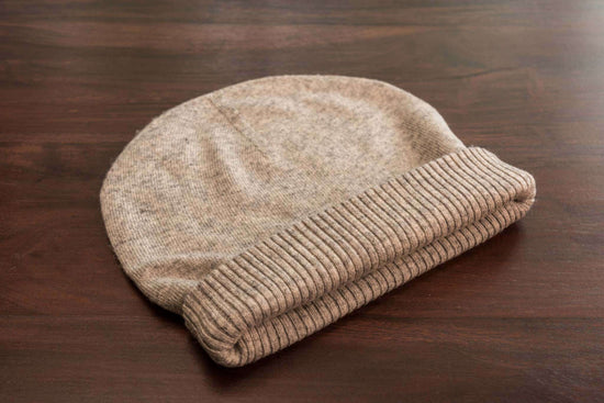 Auf dem Bild liegt eine hellgraue, gestrickte Beanie-Mütze mit glatter Oberfläche und einem breiten, gerippten Umschlag auf einer dunklen Holzoberfläche. Die Mütze ist zusammengelegt, wirkt weich und scheint aus einem warmen Material wie Kaschmir oder Wolle gefertigt zu sein. Es sind keine weiteren Objekte im Bild zu erkennen.