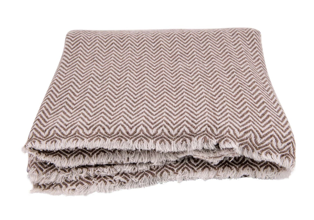 Eine zusammengefaltete Decke mit auffälligem Fischgrätenmuster in graubraunen und beigen Tönen, Fransen an den Rändern.